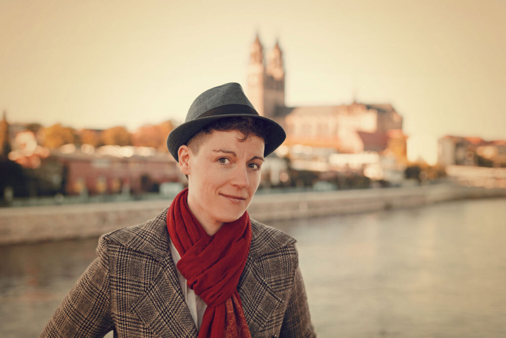 Auf dem Foto ist das Porträt der Geschäftsinhaberin Marlen Weller-Menzel zu sehen. Das Foto wurde draußen aufgenommen und zeigt Marlen Weller-Menzel vor dem Hintergrund der Stadt Magdeburg. Leicht verschwommen sieht man rechts im Hintergrund den Dom zu Magdeburg und den Fluss die Elbe.